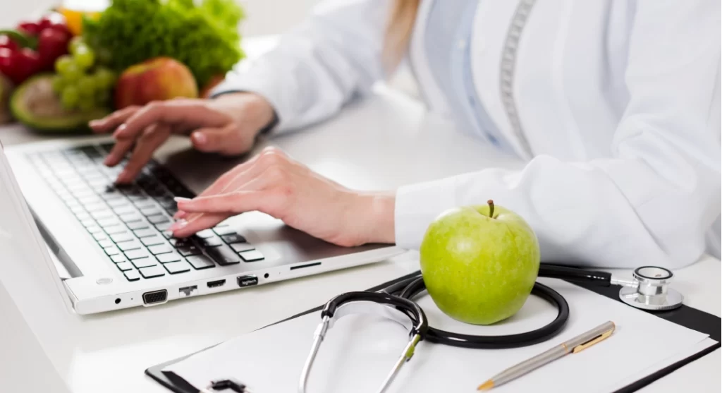 Profesional de nutrición trabajando en laptop con frutas, estetoscopio y manzana verde sobre el escritorio